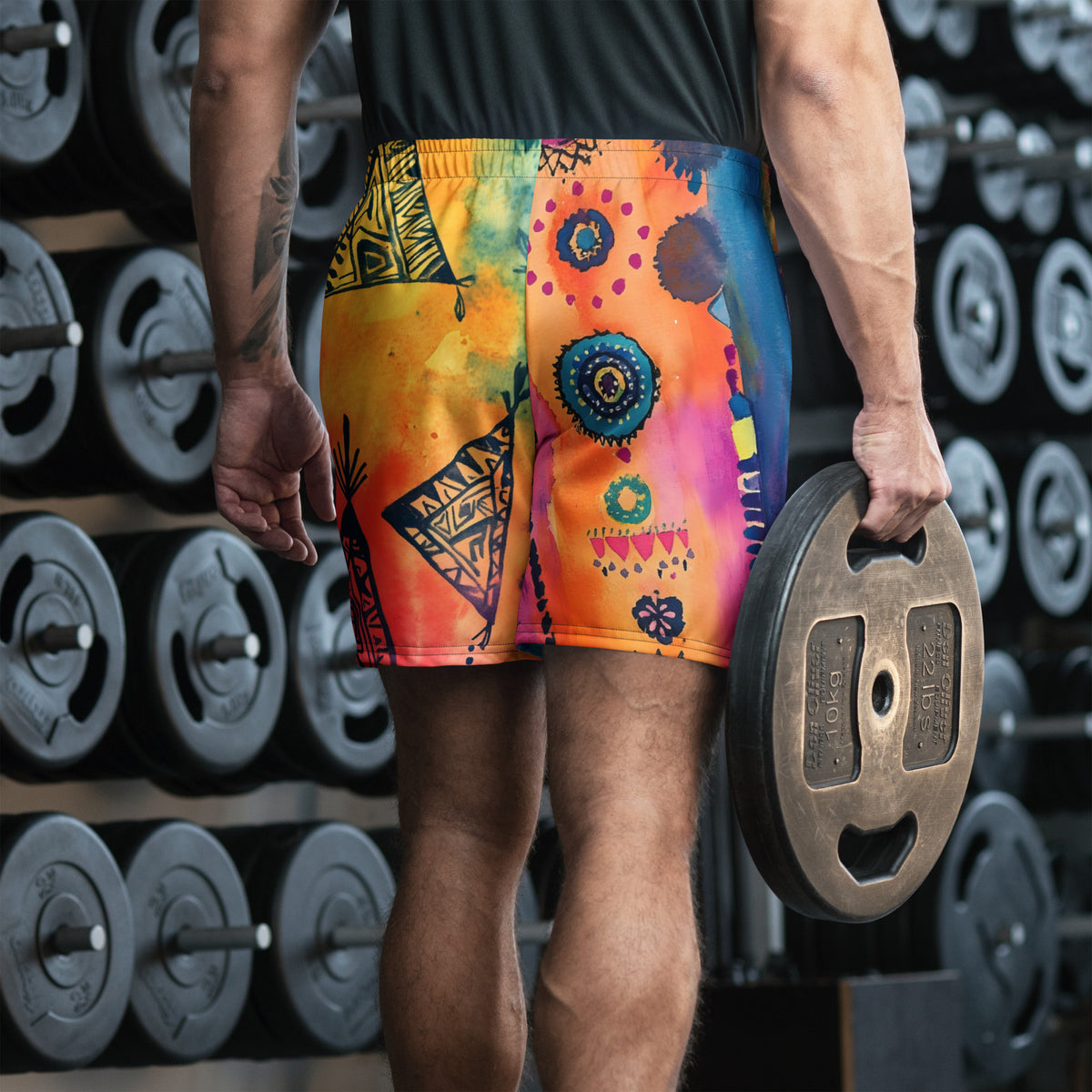 Aztec-inspired long sports shorts featuring a colorful tribal geometric abstract print with triangles, sun-like symbols, dots, decorative linework, and organic motifs in yellow, orange, coral, pink, magenta, purple, turquoise, teal, and navy. Long athletic cut with elastic waistband and white drawstring