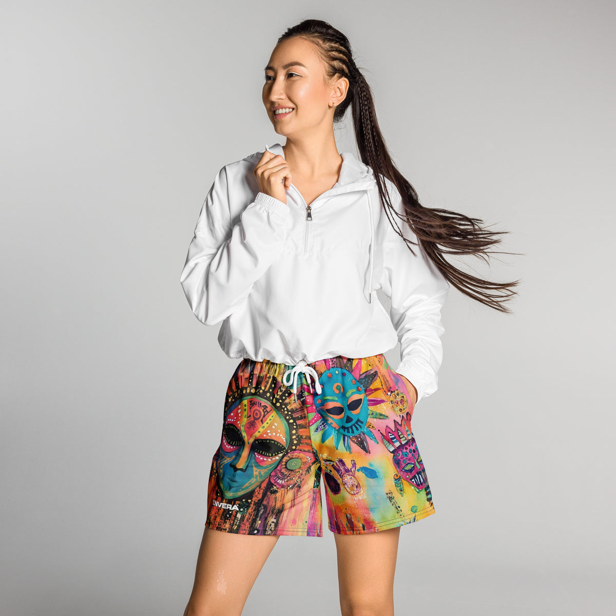 Long sports shorts featuring a vibrant psychedelic tribal mask print with expressive painted faces, decorative folk-art symbols, abstract shapes, brushstrokes, and colorful collage-like textures in turquoise, teal, coral, pink, magenta, orange, yellow, purple, black, and earthy tones. Unisex athletic design with a longer cut, elastic waistband, and bold artistic all-over pattern
