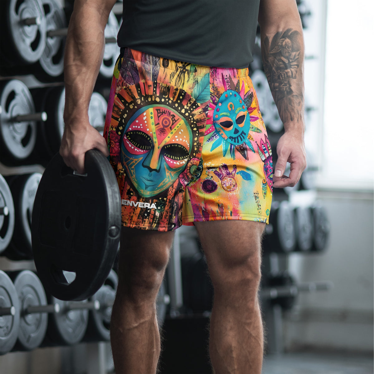 Long sports shorts featuring a vibrant psychedelic tribal mask print with expressive painted faces, decorative folk-art symbols, abstract shapes, brushstrokes, and colorful collage-like textures in turquoise, teal, coral, pink, magenta, orange, yellow, purple, black, and earthy tones. Unisex athletic design with a longer cut, elastic waistband, and bold artistic all-over pattern