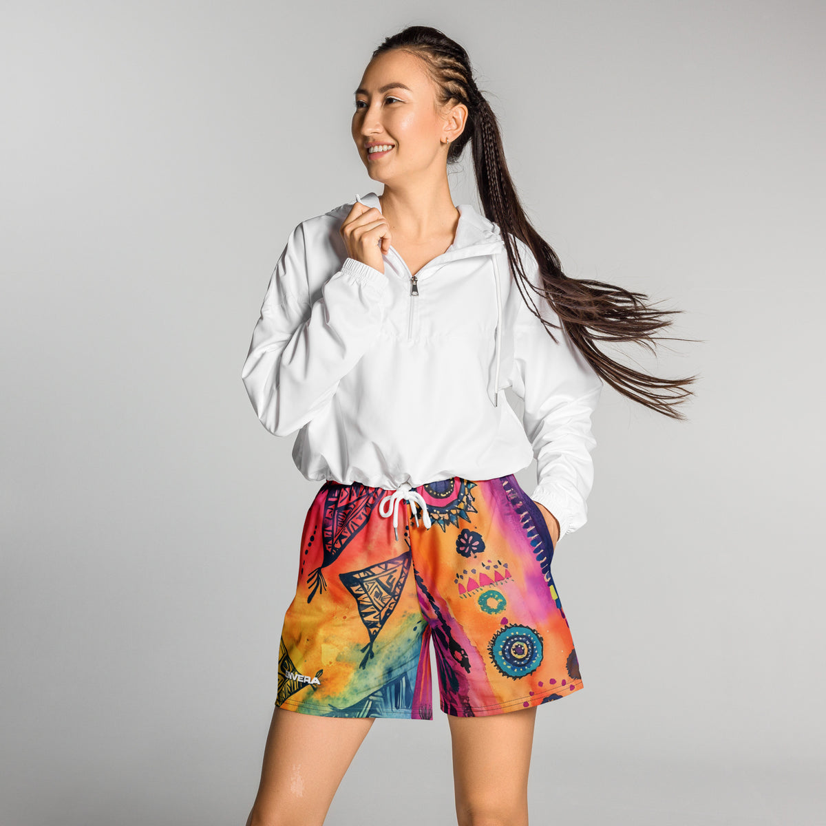 Aztec-inspired long sports shorts featuring a colorful tribal geometric abstract print with triangles, sun-like symbols, dots, decorative linework, and organic motifs in yellow, orange, coral, pink, magenta, purple, turquoise, teal, and navy. Long athletic cut with elastic waistband and white drawstring