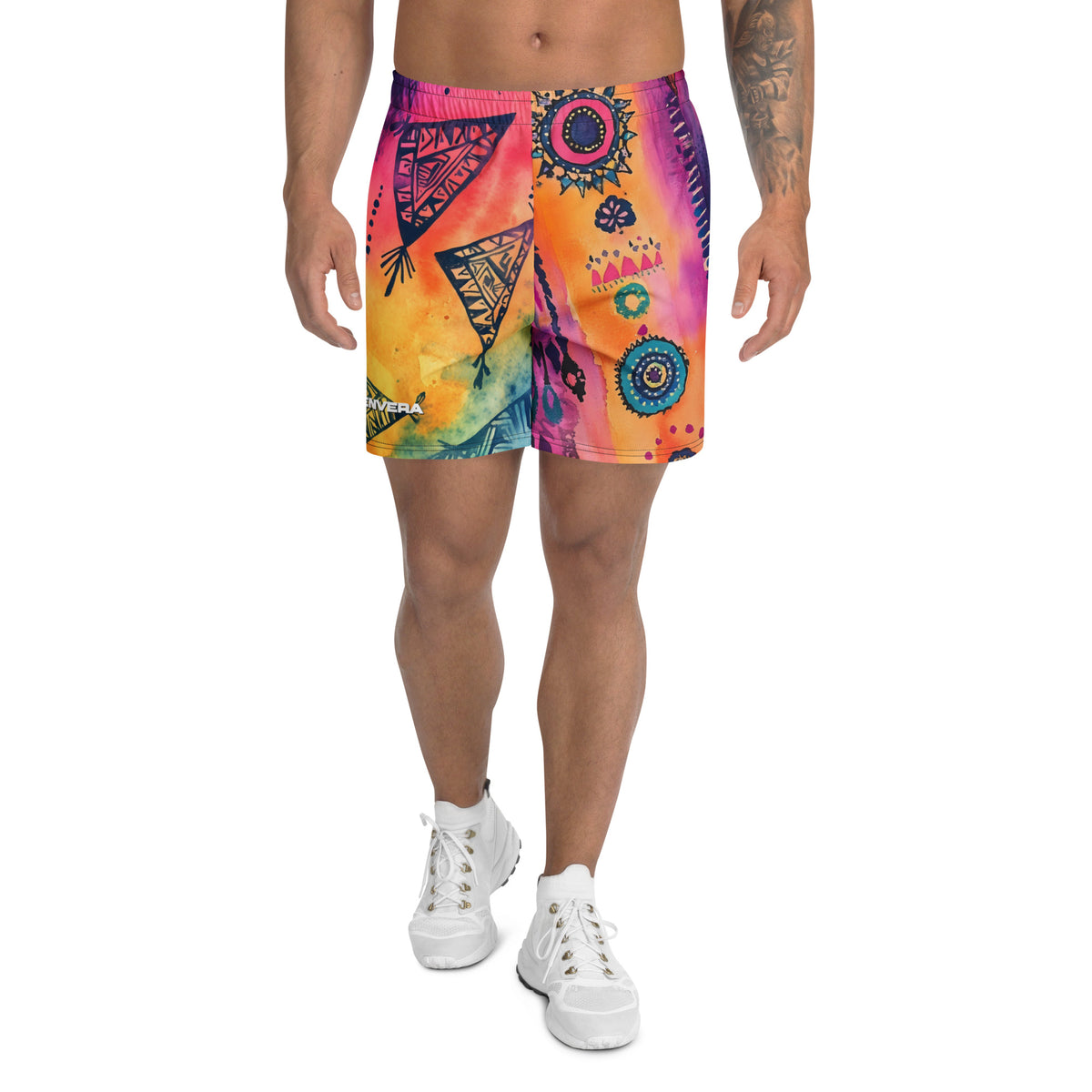 Aztec-inspired long sports shorts featuring a colorful tribal geometric abstract print with triangles, sun-like symbols, dots, decorative linework, and organic motifs in yellow, orange, coral, pink, magenta, purple, turquoise, teal, and navy. Long athletic cut with elastic waistband and white drawstring