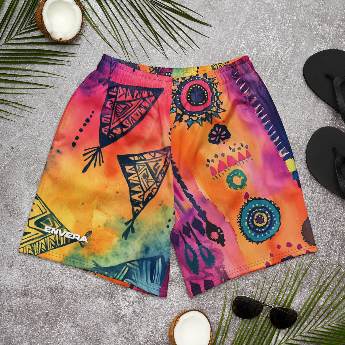 Aztec-inspired long sports shorts featuring a colorful tribal geometric abstract print with triangles, sun-like symbols, dots, decorative linework, and organic motifs in yellow, orange, coral, pink, magenta, purple, turquoise, teal, and navy. Long athletic cut with elastic waistband and white drawstring