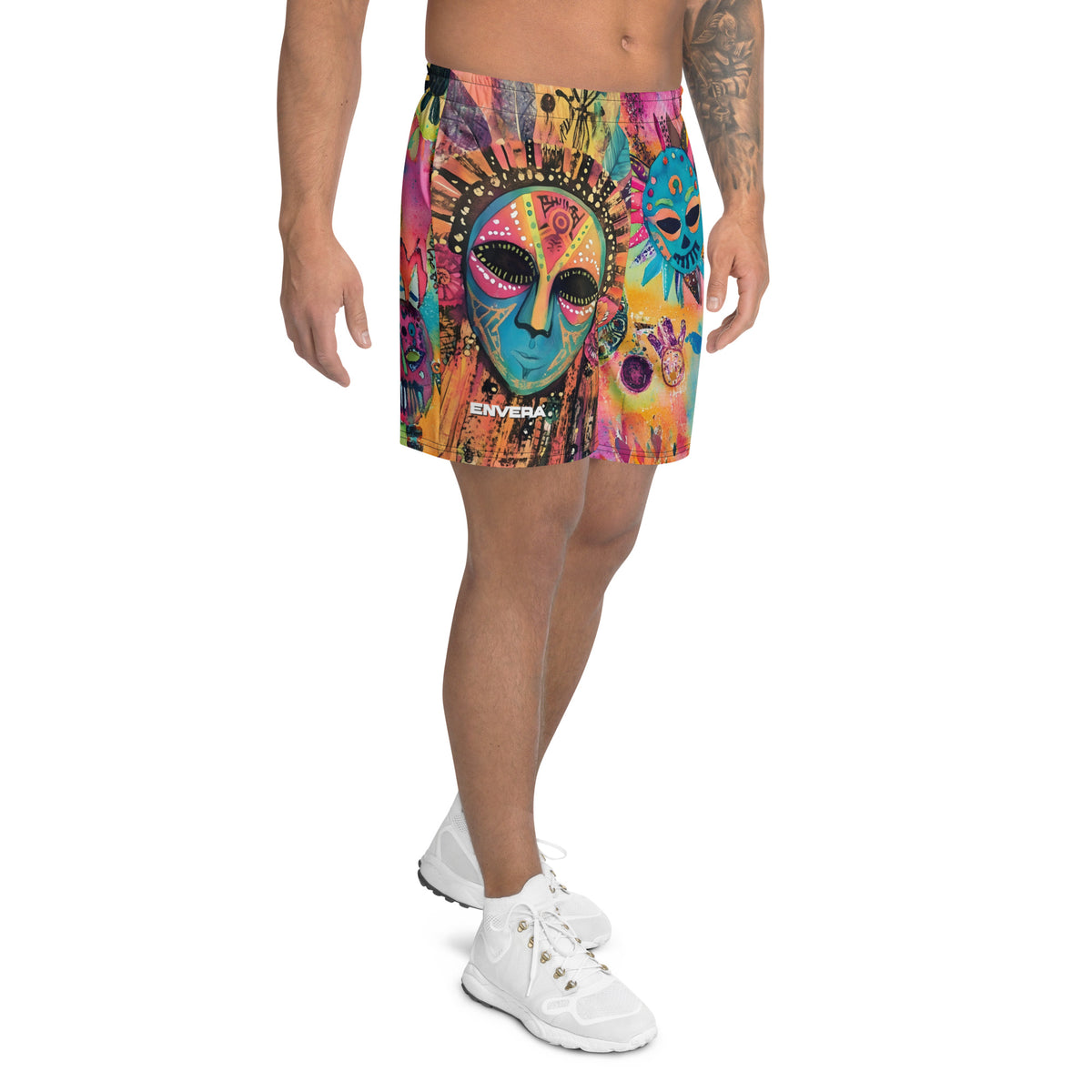 Long sports shorts featuring a vibrant psychedelic tribal mask print with expressive painted faces, decorative folk-art symbols, abstract shapes, brushstrokes, and colorful collage-like textures in turquoise, teal, coral, pink, magenta, orange, yellow, purple, black, and earthy tones. Unisex athletic design with a longer cut, elastic waistband, and bold artistic all-over pattern